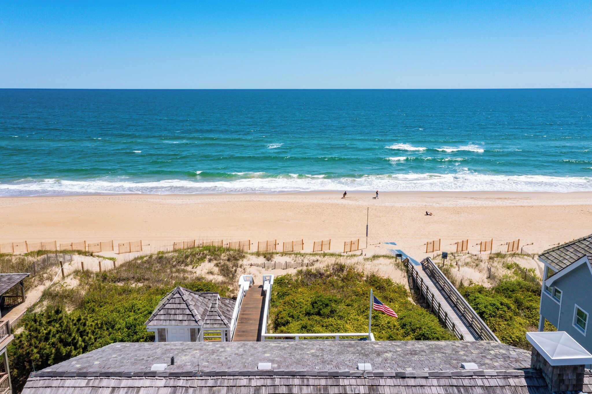A beach view with a wooden boardwalk leading to a house with a flag.