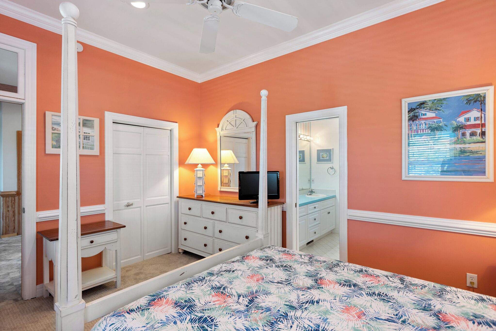 A bedroom with an orange wall, white furniture, a bed with a floral bedspread, a dresser, a vanity with a mirror, and a ceiling fan.