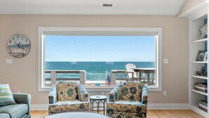 A living room with a large window overlooking the ocean, two floral armchairs, a small round table, and a clock on the wall.