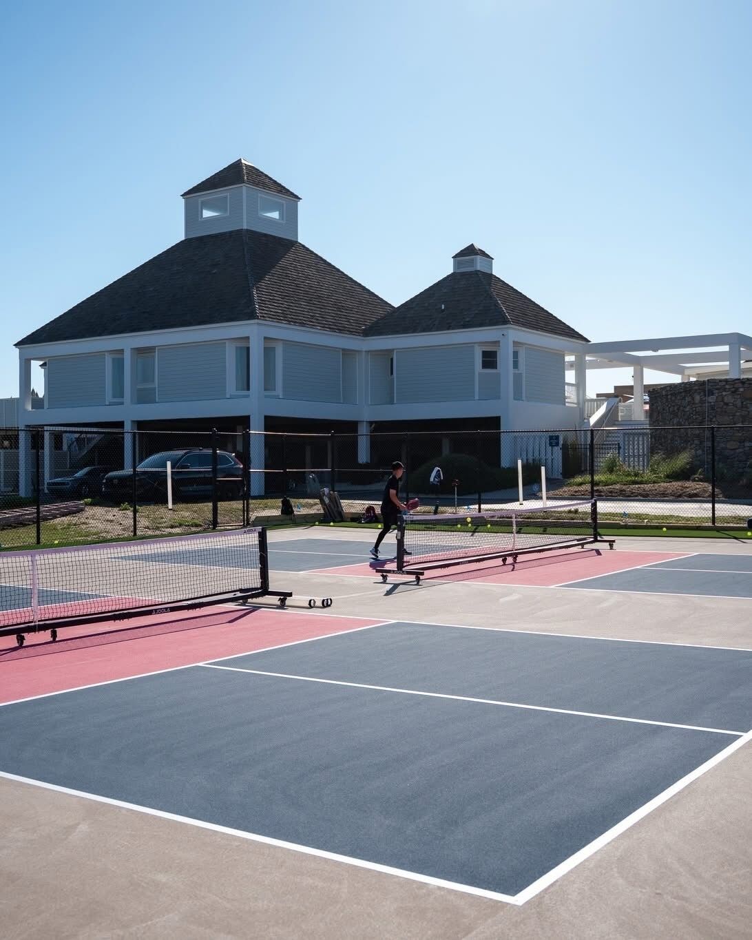 Een tennisbaan met een net, een persoon aan het spelen, en een gebouw met een garage op de achtergrond.