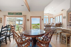 Un coin repas avec une table ronde et des chaises, une vue sur la mer par une porte vitrée, et un espace bar avec des meubles en osier.