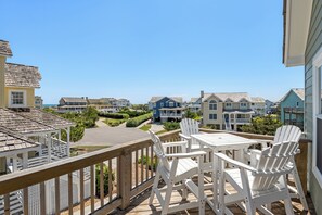 Une terrasse en bois avec des chaises blanches et une table, d’où l’on a vue sur une rangée de maisons de plage.