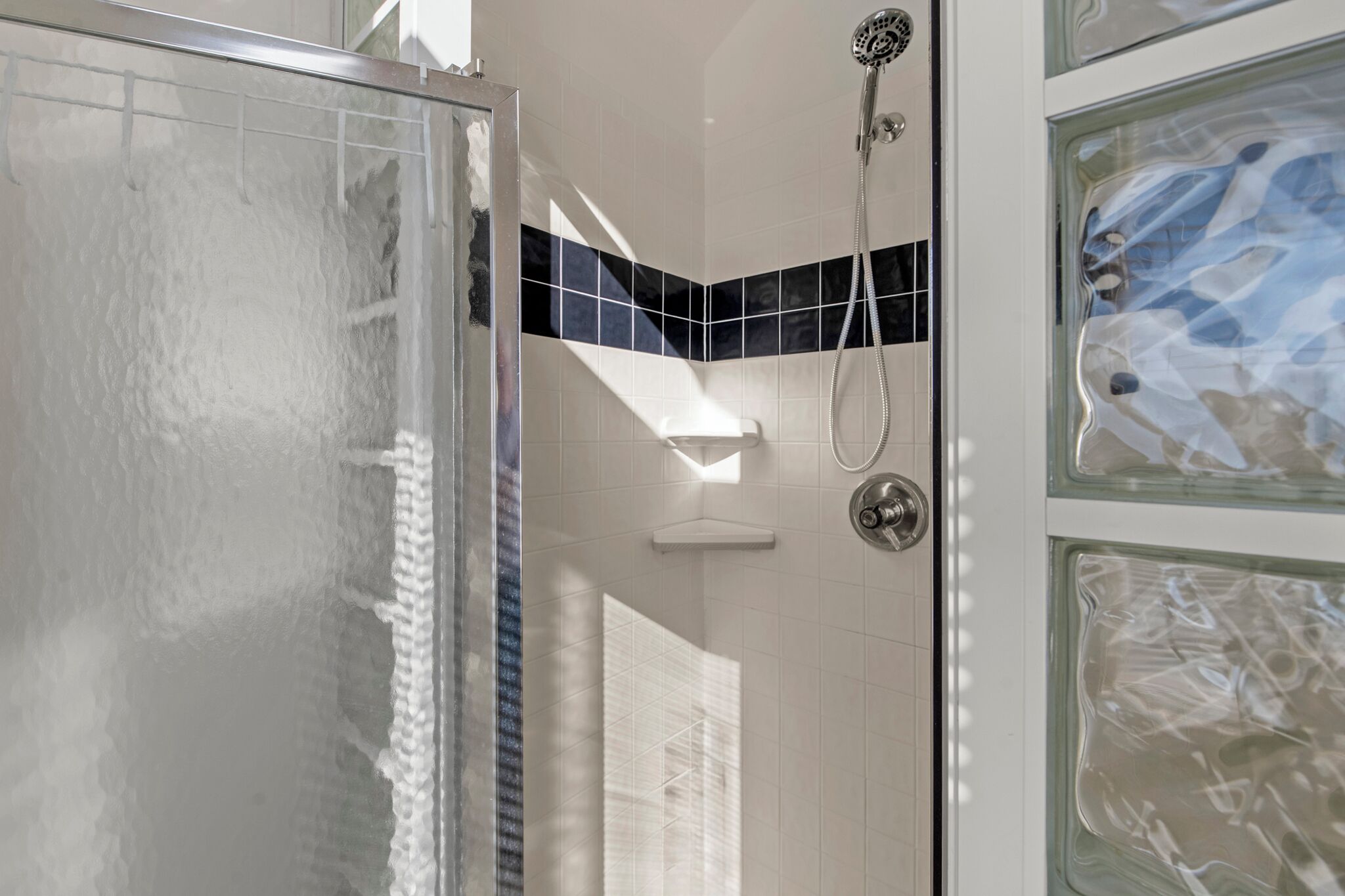 A shower area with a glass door, a handheld showerhead, and a corner tile design.