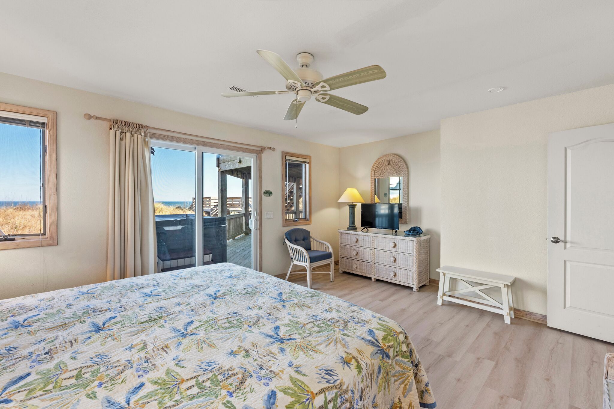 A bedroom with a large bed, a ceiling fan, a dresser, a chair, a bench, a TV, and a balcony with a view.