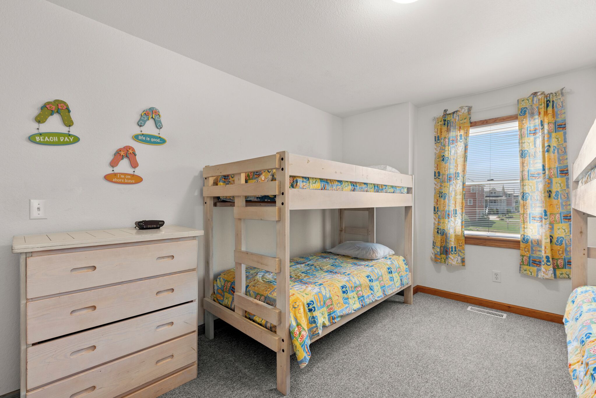 A bunk bed with a wooden frame and a colorful bedspread, a wooden dresser with drawers, and a window with patterned curtains.