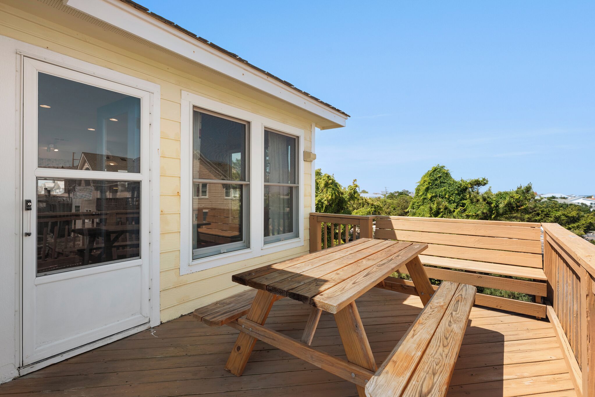 A wooden deck with a picnic table and benches, a white door, and large windows.