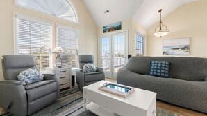 A living room with a gray sofa, two gray recliners, a white coffee table, and a large window with blinds. - A Change In Latitude: Pet-friendly! Shared Pool, Private Hot Tub (Nags Head)