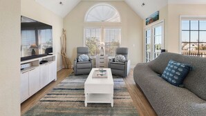 A living room with a white coffee table, grey armchairs, a grey sofa, and a TV on a white cabinet. - A Change In Latitude: Pet-friendly! Shared Pool, Private Hot Tub (Nags Head)