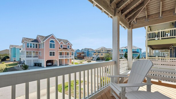 A wooden deck with white chairs and a view of colorful houses. - A Change In Latitude: Pet-friendly! Shared Pool, Private Hot Tub (Nags Head)