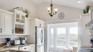 A kitchen with white cabinets, a stainless steel refrigerator, a coffee maker, and a window overlooking a landscape. - Hook, Wine And Sinker: Private Pool, Hot Tub, Free Community Amenities! (Corolla)