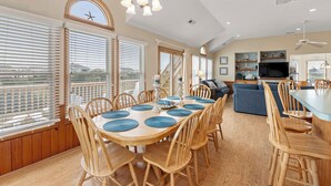 A spacious dining area with a large wooden table and chairs, a chandelier, and large windows with blinds. - Ebbtide: Private Pool, Screened Porch, Ocean Views (Corolla)