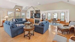 A spacious living room with a fireplace, blue sofas, a television, and wicker furniture. - Ebbtide: Private Pool, Screened Porch, Ocean Views (Corolla)