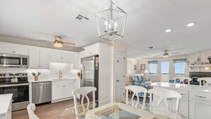 Una cocina moderna con armarios blancos, electrodomésticos de acero inoxidable, una mesa de comedor de vidrio y un ventilador de techo.