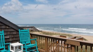 Terraza de madera con sillas turquesa y una pequeña mesa con vista a una playa y olas.