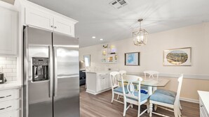 Una cocina moderna con un refrigerador de acero inoxidable, armarios blancos, una zona de comedor con una mesa de vidrio y sillas blancas, y una lámpara colgante.