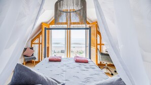 A bedroom with a bed, pillows, a chair, a desk, and a view of the beach through a large window.