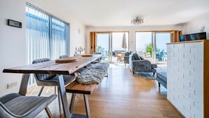 A modern living room with a wooden dining table, grey sofas, and a view of the sea through large windows.