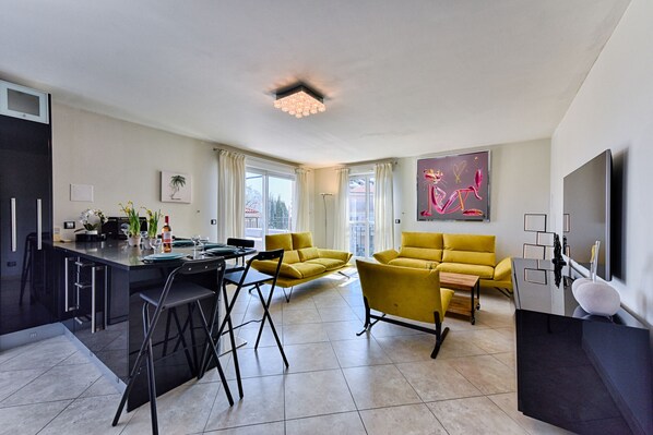 A modern living room with a yellow sofa, a dining area with a bar, and a large TV on the wall.
