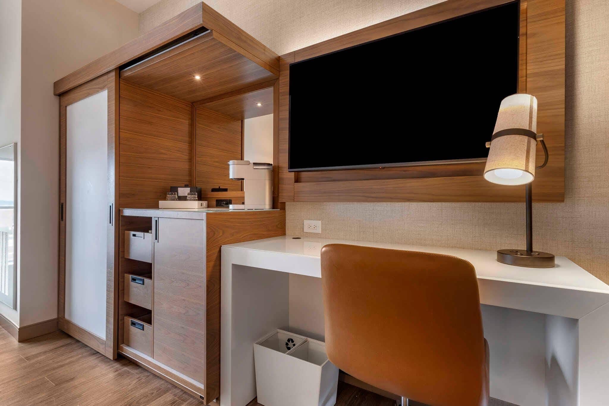 A modern bedroom with a wooden wardrobe, a desk with a lamp, and a large TV mounted on the wall.