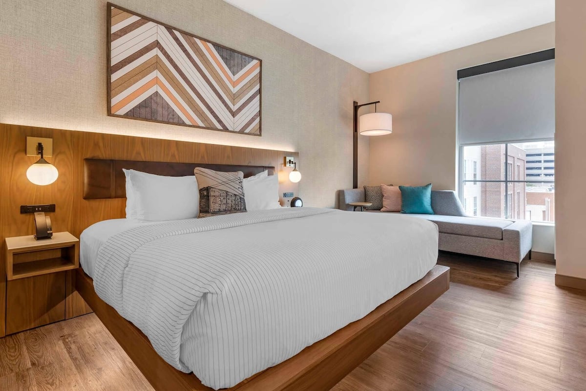 A modern hotel room with a large bed, wooden headboard, bedside lamps, a wall-mounted artwork, a sofa, and a large window with a view of buildings.