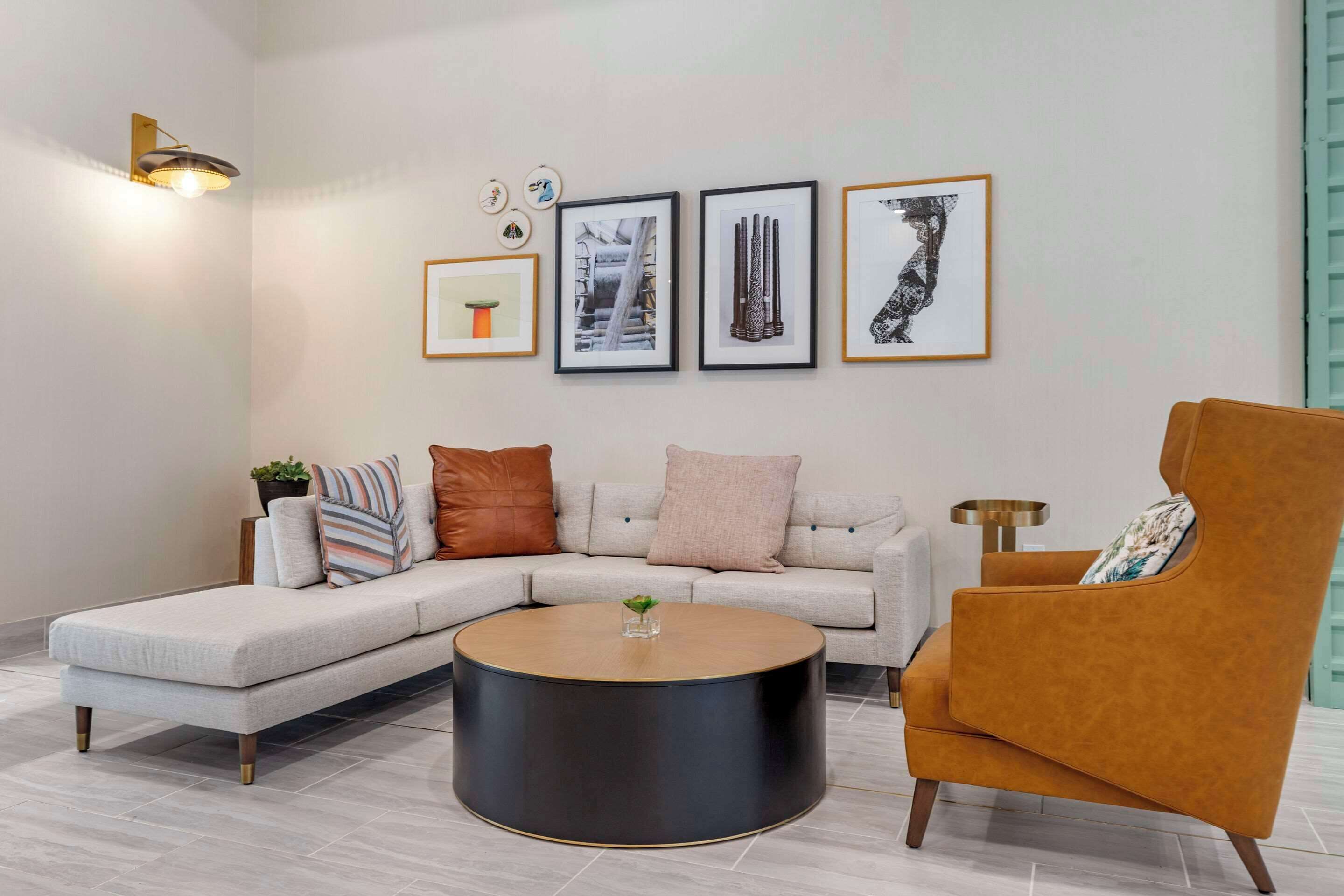 A modern living room with a sectional sofa, a round coffee table, and a single armchair. There are framed artworks on the wall and a floor lamp.