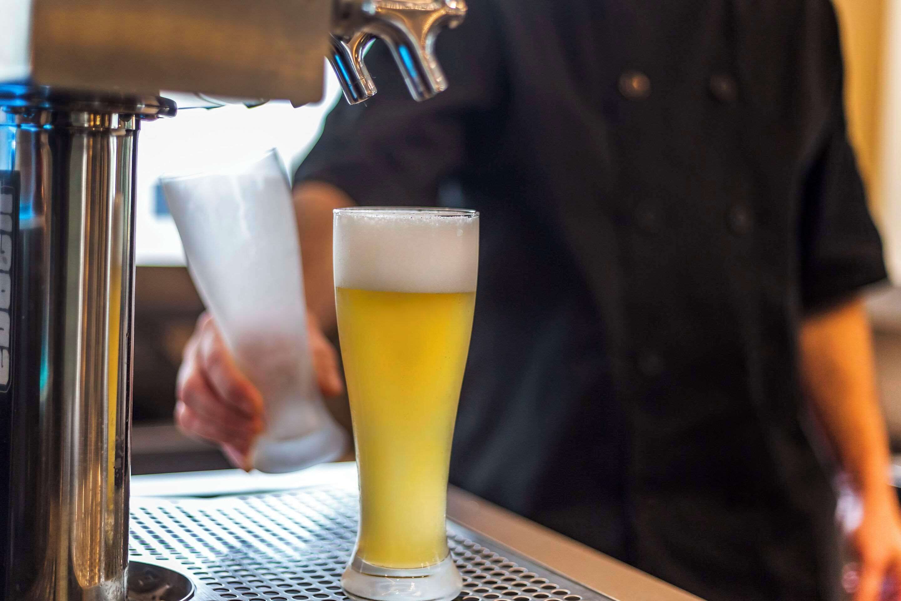 Een hoog glas bier aan een bar, met een biertappunt en een barista op de achtergrond.