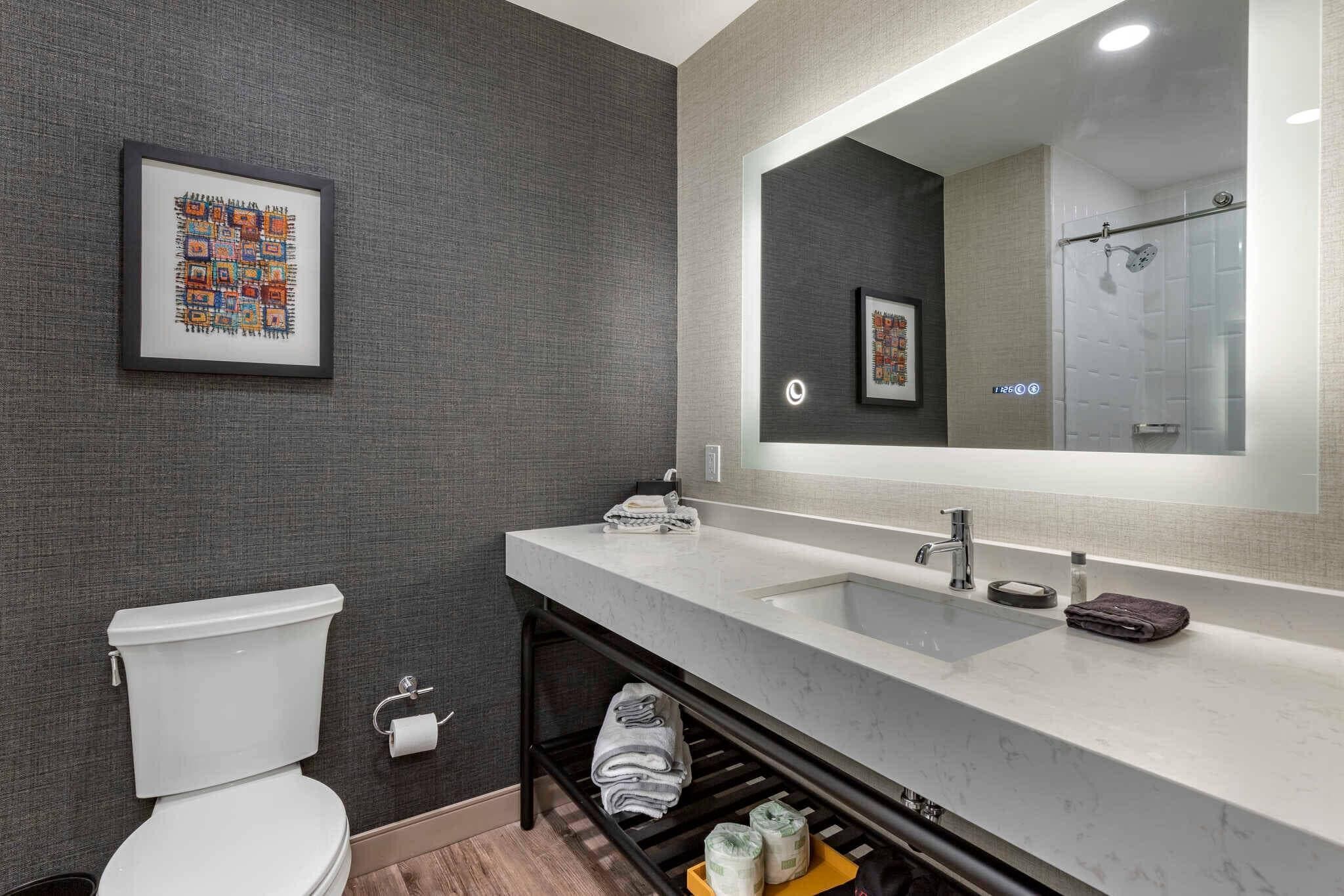 A modern bathroom with a toilet, sink, and shower. There is a framed artwork on the wall and a towel rack below the sink.