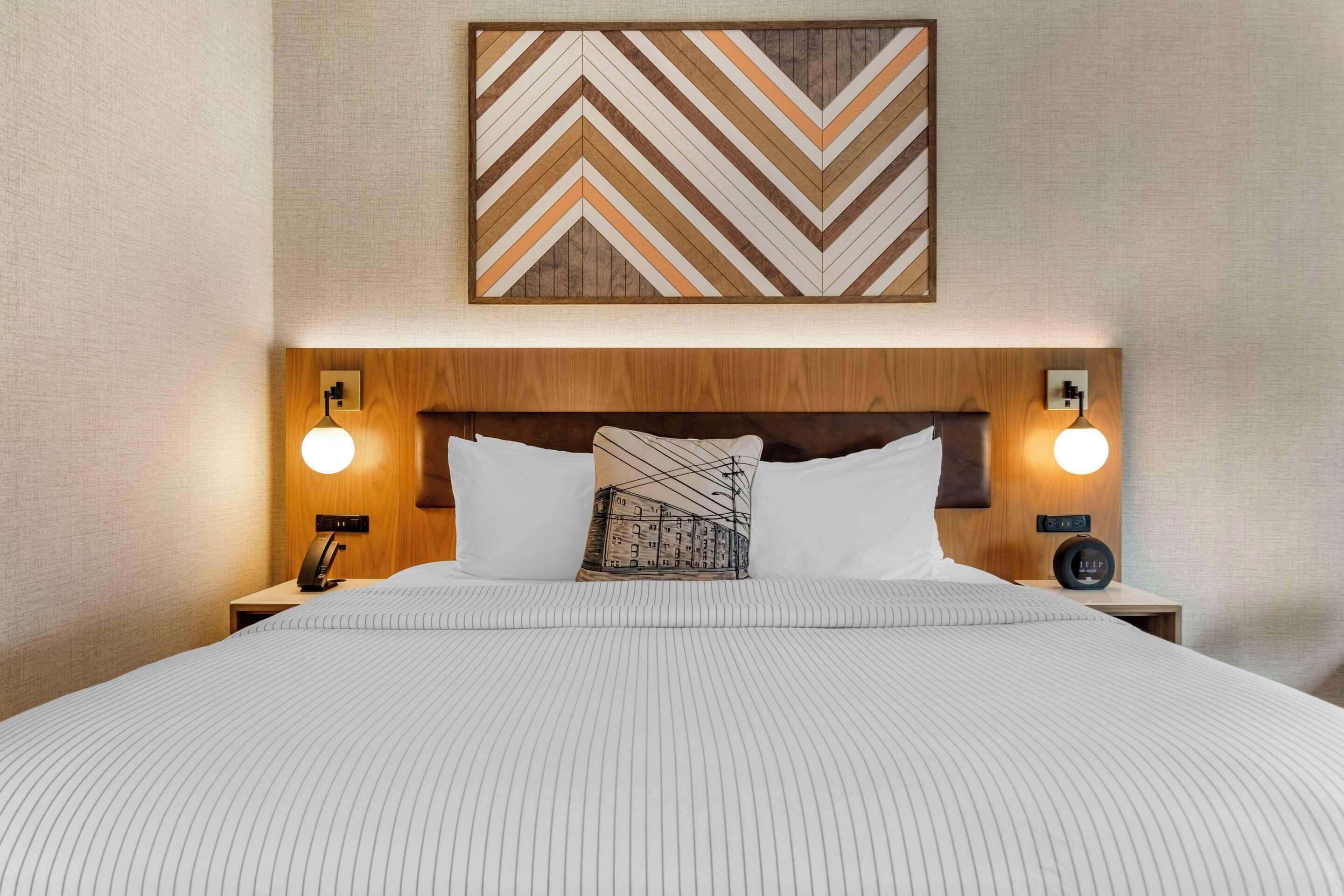 A neatly made bed with a striped bedspread, two bedside tables with lamps, a wooden headboard, and a chevron-patterned artwork above the bed.