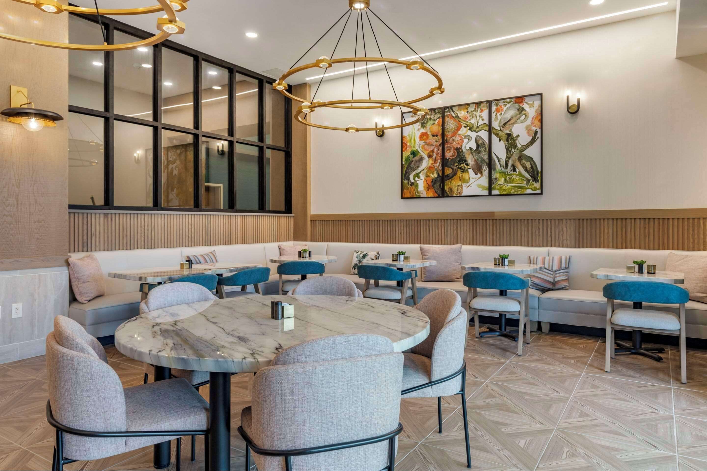 A modern dining area with a round marble table, cushioned seating, and a large window with blinds.