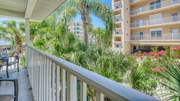 A balcony with a railing, surrounded by palm trees and pink flowers, overlooking a multi-story building. - You and Me and the Sea: 2  BR, 2  BA Condominium in Indian Rocks Beach, Sleeps 4 (Indian Rocks Beach)