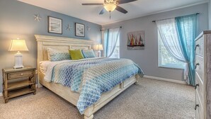 Un dormitorio con cama, ventilador de techo, mesita de noche, cómoda y ventana con cortinas.