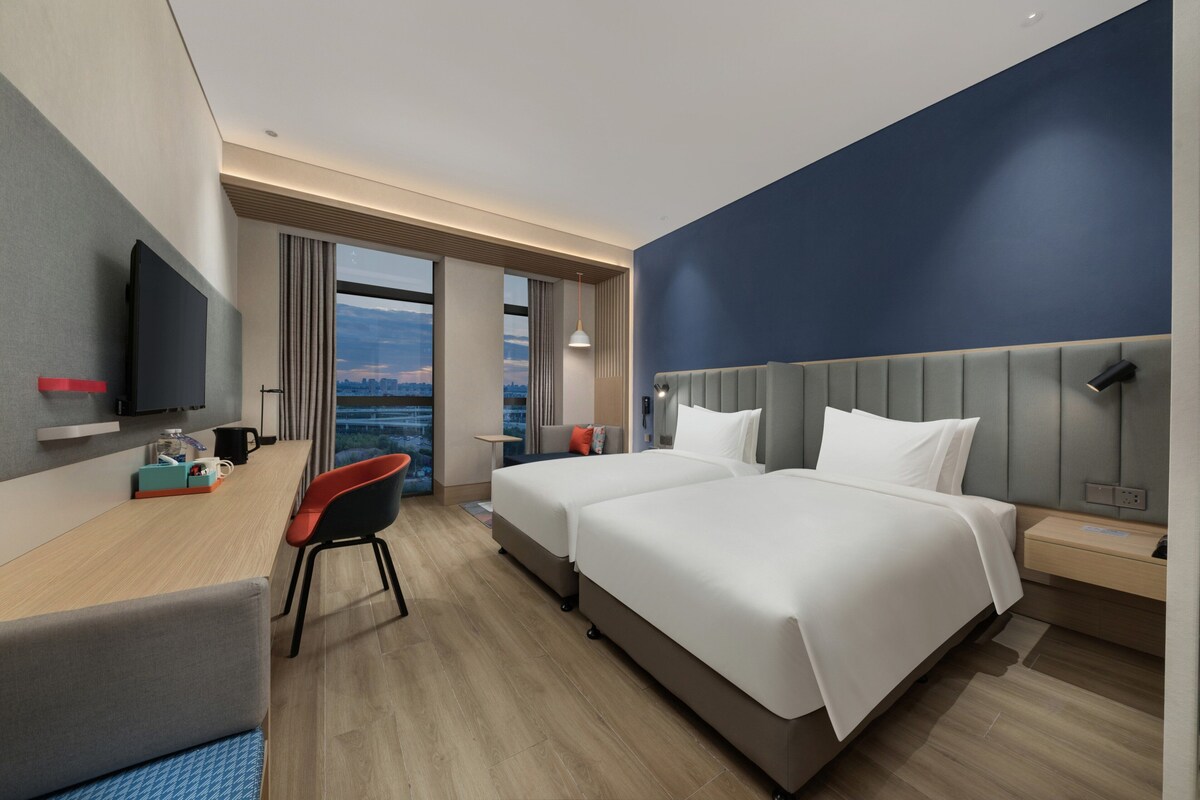 Ein modernes Hotelzimmer mit einem großen Bett, einem Schreibtisch mit Stuhl, einem Fernseher und Blick auf die Stadtlandschaft.