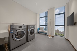 Ein moderner Waschraum mit zwei Frontlader-Waschmaschinen, einer Sitzbank, einem großen Fenster mit Blick auf die Stadt und einem an der Wand befestigten Fernseher.