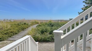 A wooden deck leading to a white staircase with a clear view of the beach and dunes in the background.