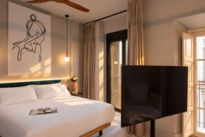 A hotel room with a bed, a flat-screen TV, a wall art piece, a window with curtains, and a bedside table with a vase and a bottle. - Coeo Peña Design Apartments (Málaga)