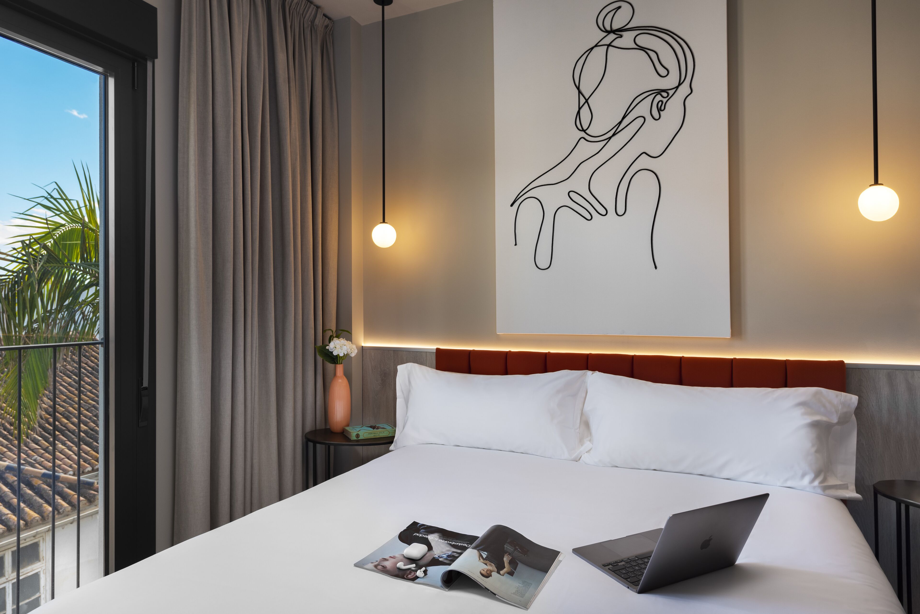 Une chambre d’hôtel moderne avec un grand lit, un ordinateur portable posé sur le lit, un magazine et une œuvre d’art murale.