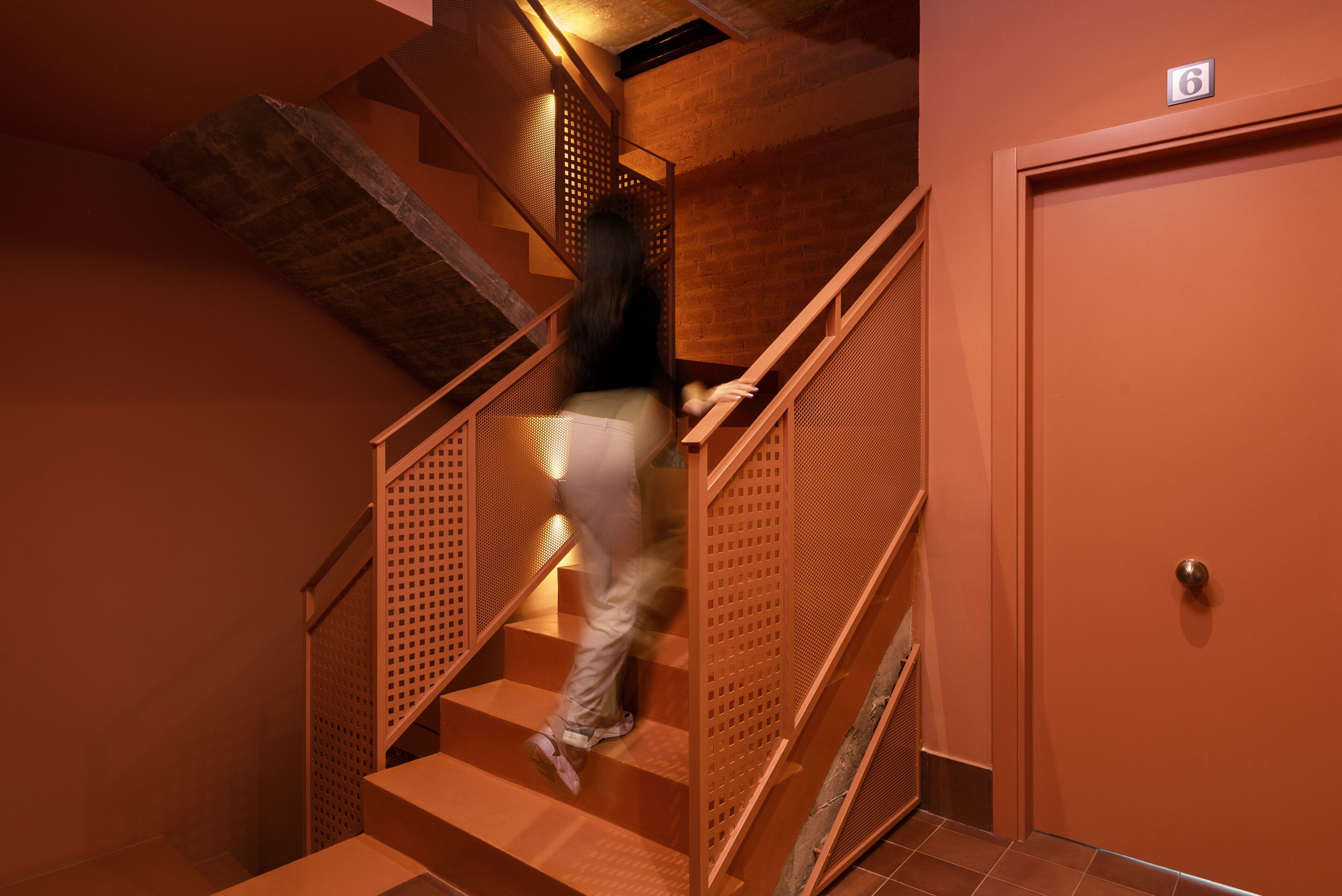 Una persona sube una escalera de madera con barandilla de metal, que conduce a una puerta con el número 6.