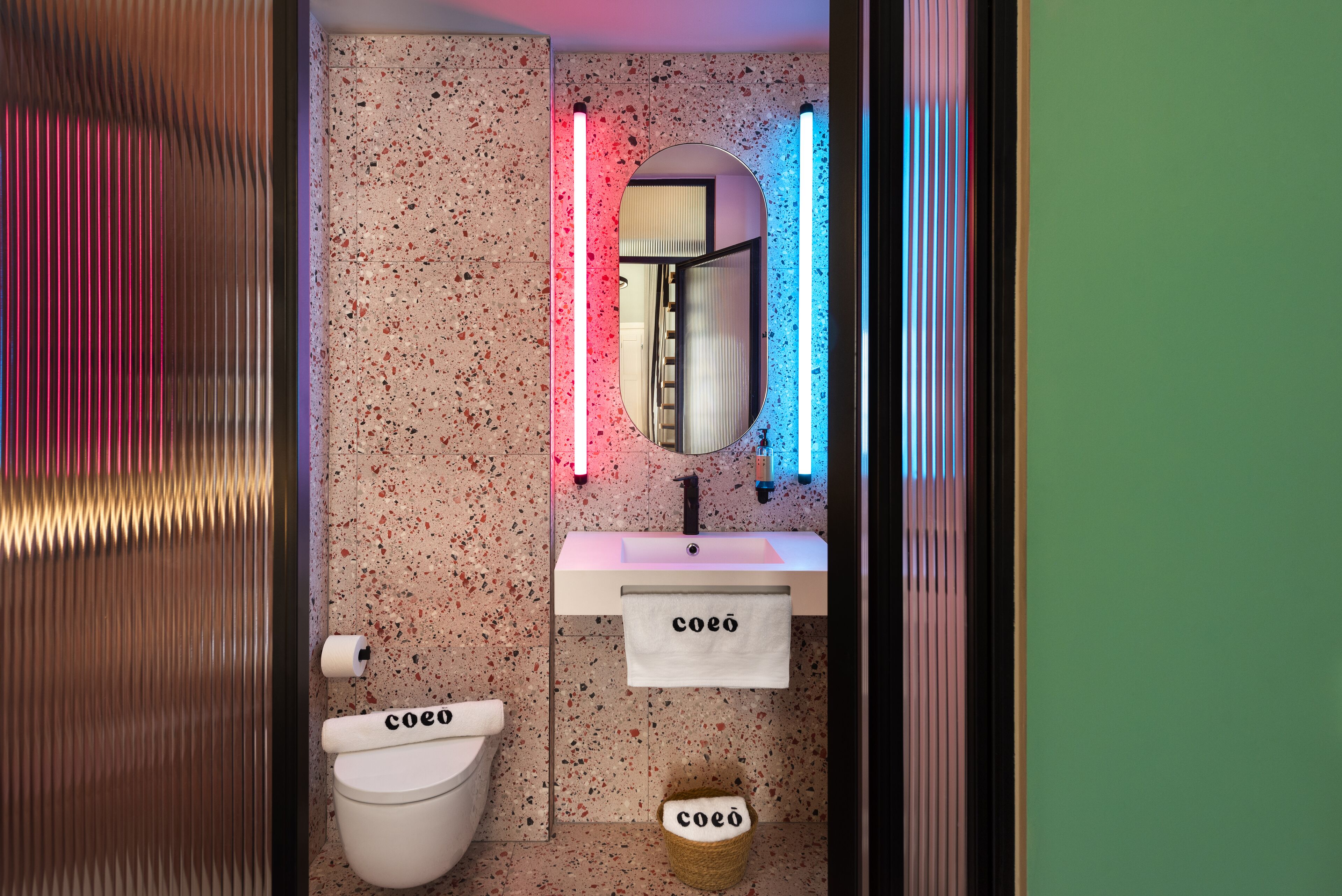 Ein Badezimmer mit Toilette, Waschbecken und Spiegel. Die Wände haben ein gemustertes Dekor, und über dem Spiegel befinden sich zwei vertikale Lichtleisten.