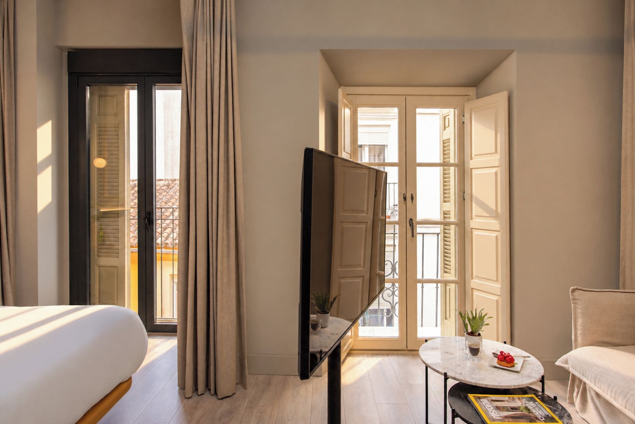 A hotel room with a bed, a TV, a coffee table, and a balcony with curtains.