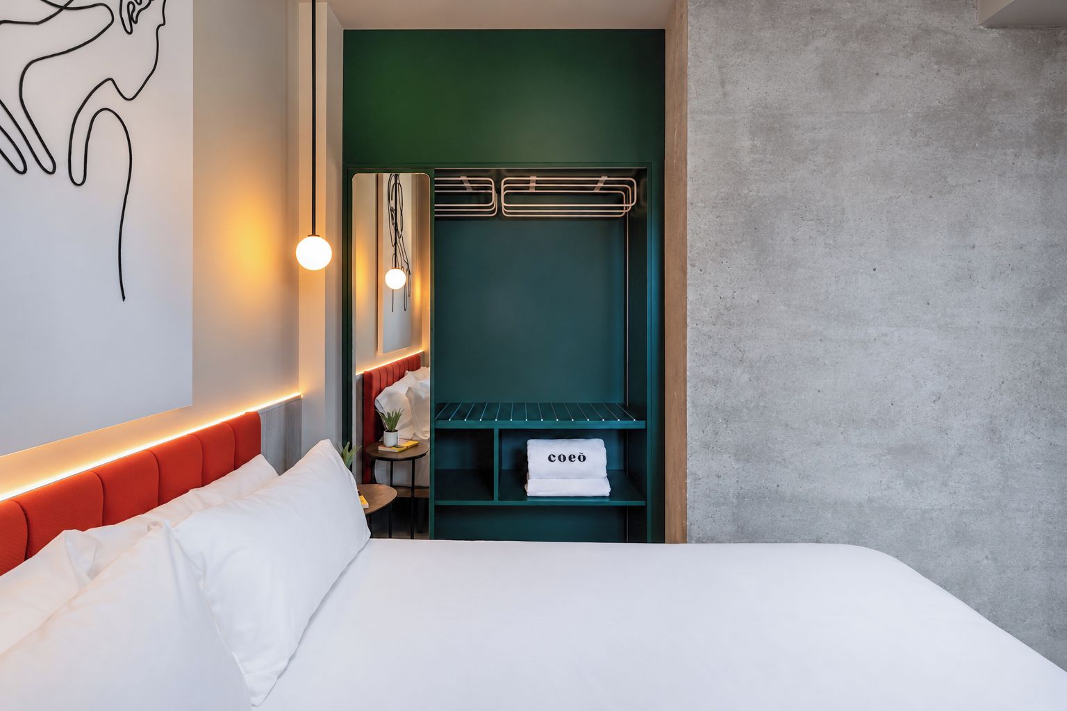 A hotel room with a bed, a green wall, a coat rack, and a towel rack with a towel labeled 'coeo'.
