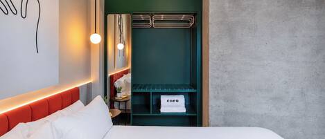 A hotel room with a bed, a green wall, a coat rack, and a towel rack with a towel labeled 'coeo'.