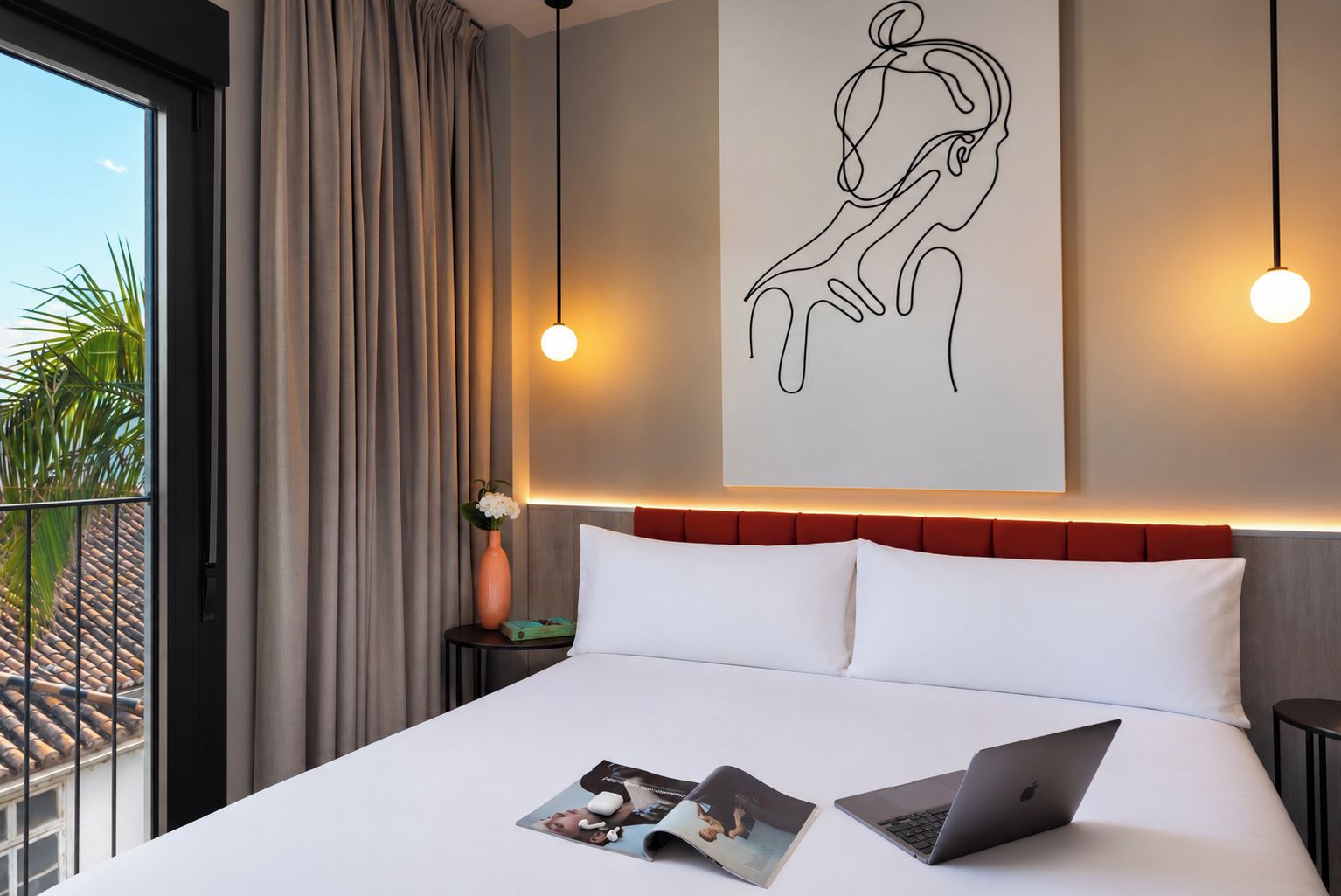 A hotel room with a large bed, a laptop, a magazine, a vase with flowers, and a wall art piece.