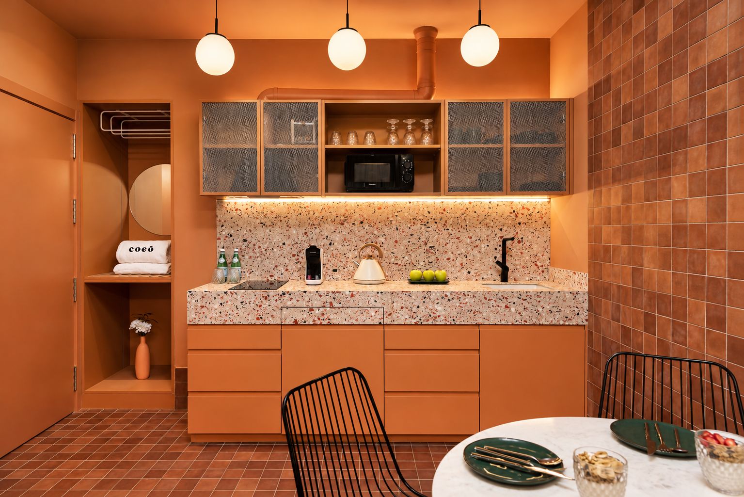 A compact kitchen with a granite countertop, a microwave, and a sink. There are cabinets above the counter, a chair, and a dining table with a plate and cutlery.