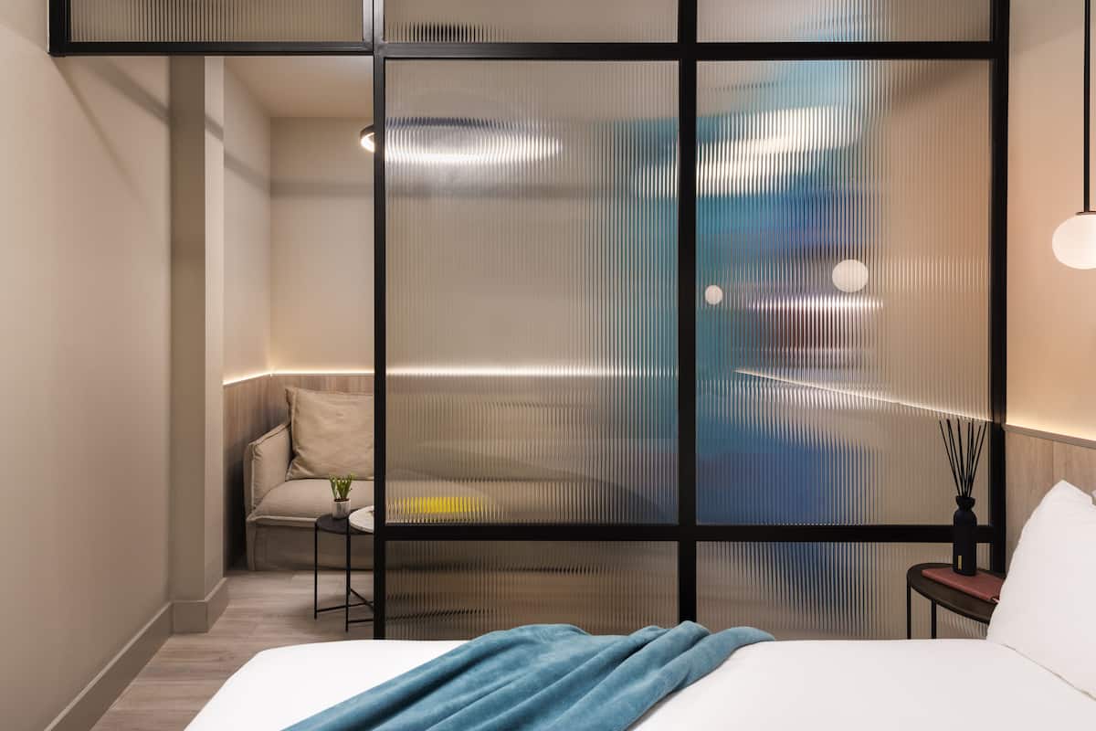 Quarto de hotel moderno com uma cama grande, um pequeno espaço de estar com uma cadeira e uma mesa, e um divisória decorativa com linhas verticais.