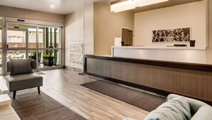A modern reception area with a reception desk, a grey sofa, and a large window with a view of the outside. - MainStay Suites Dallas Northwest - Irving (Dallas)