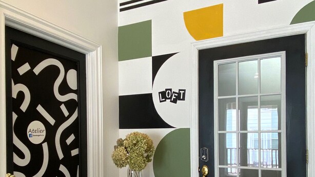 A hallway with a black door featuring a white abstract design, a round wooden table with a vase, and a large window with a view of the outside.