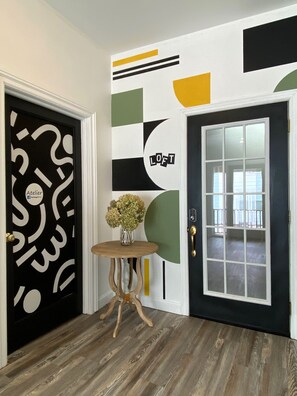 A hallway with a black door featuring a white abstract design, a round wooden table with a vase, and a large window with a view of the outside.