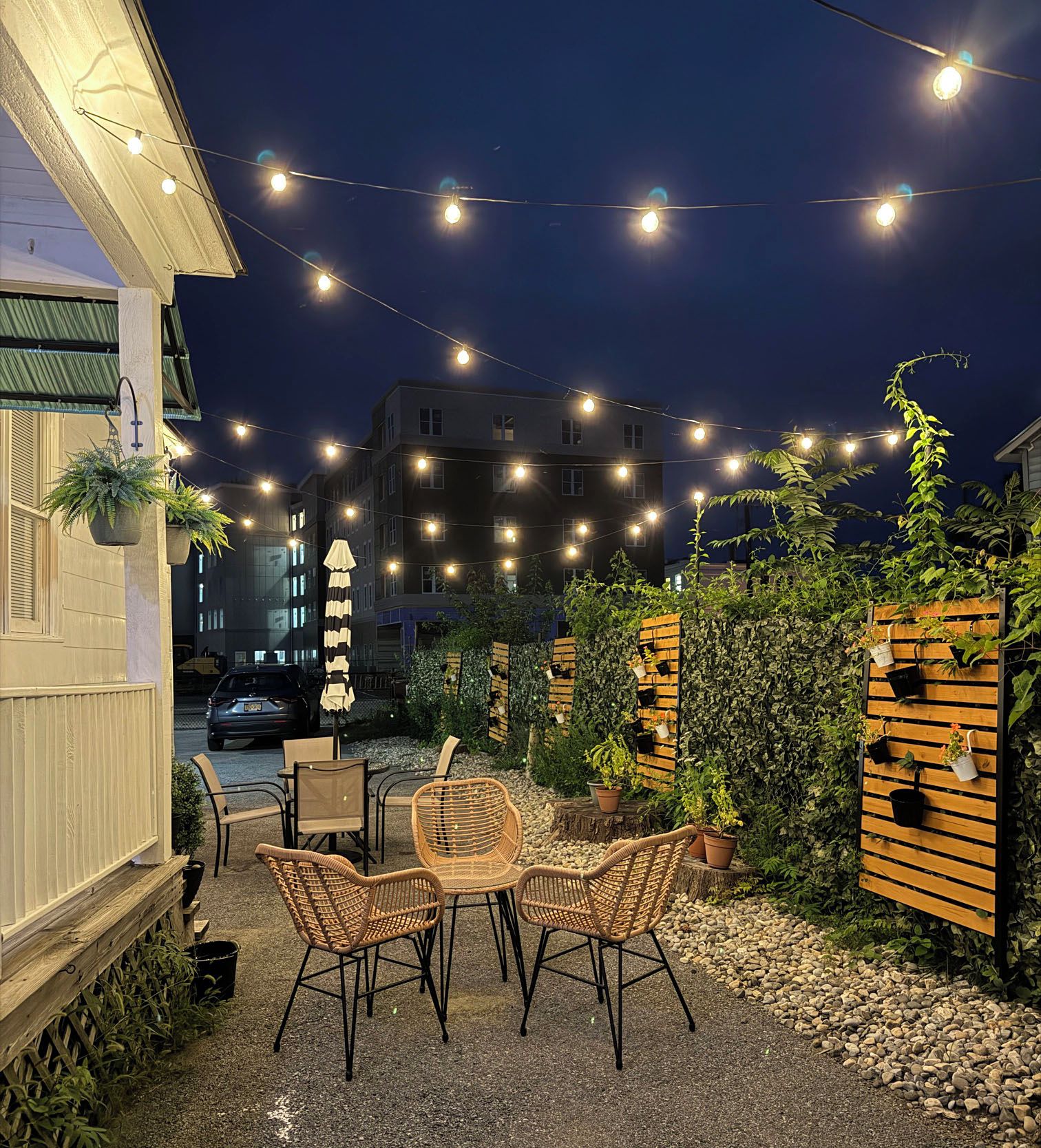 Un espacio al aire libre con sillas y mesas de mimbre, luces colgantes y plantas en macetas.