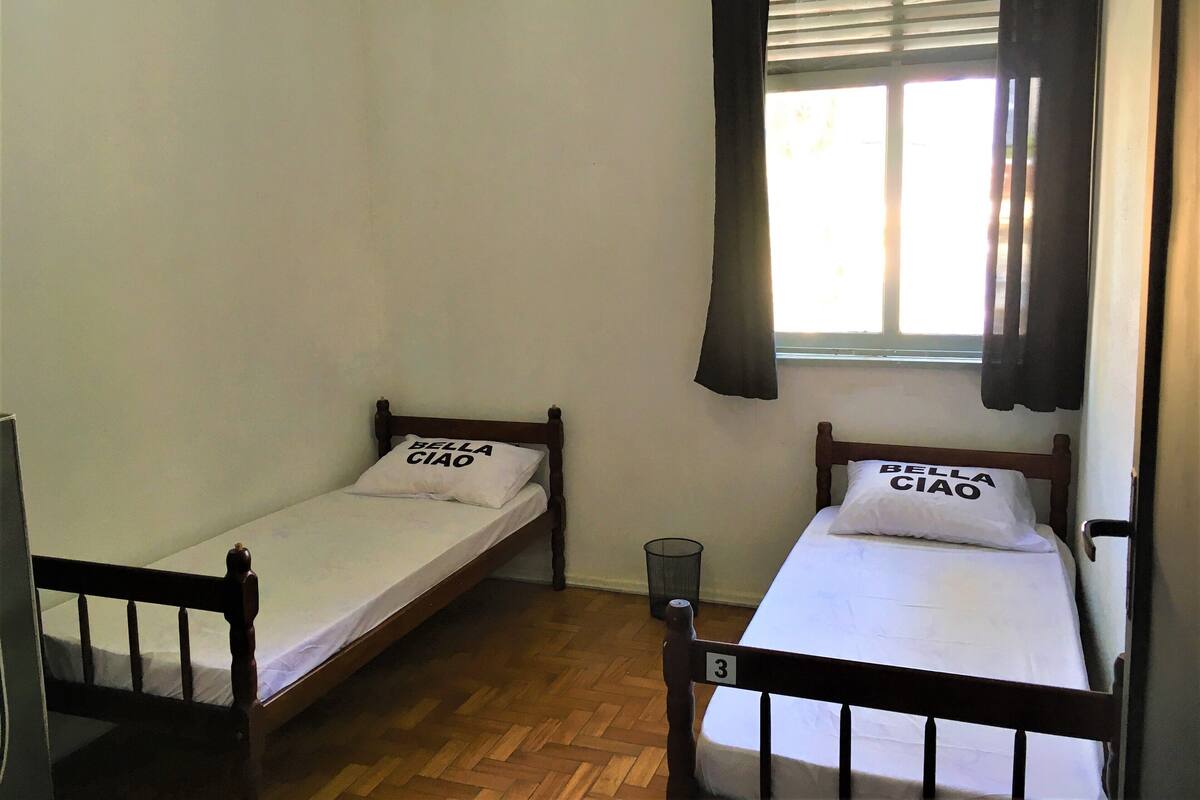 Two single beds with wooden frames and white bedding, each with a pillow labeled "Bella Ciao". A window with blinds and a curtain is visible, and a trash bin is placed against the wall.