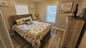 Une chambre à coucher comprenant un lit, une table de chevet, une commode et deux fenêtres équipées de stores.